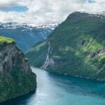 Steep green cliffs with a cascading waterfall overlooking a turquoise fjord, surrounded by snow-capped mountains under a cloudy sky.