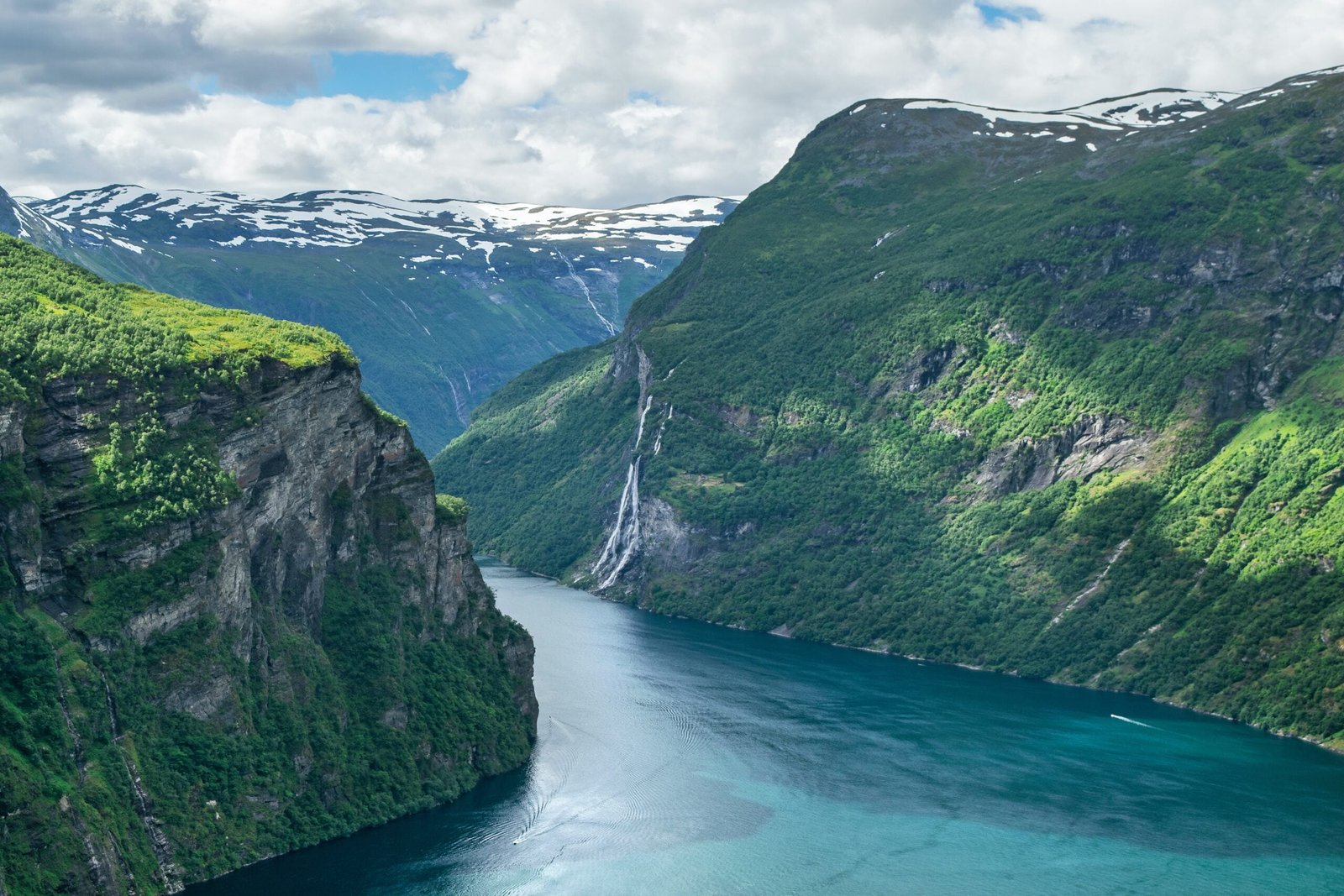 Steep green cliffs with a cascading waterfall overlooking a turquoise fjord, surrounded by snow-capped mountains under a cloudy sky.