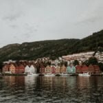 Bergen cityscape with colorful wooden houses lining the harbor, surrounded by verdant mountains under a cloud-filled sky.