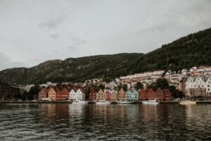 Bergen cityscape with colorful wooden houses lining the harbor, surrounded by verdant mountains under a cloud-filled sky.