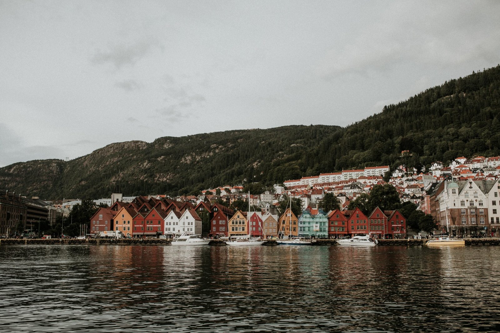 Bergen cityscape with colorful wooden houses lining the harbor, surrounded by verdant mountains under a cloud-filled sky.