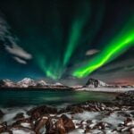 Vibrant green northern lights dancing over snowy mountain peaks, with rocky coastline and calm ocean waters in the foreground under a starry night sky.