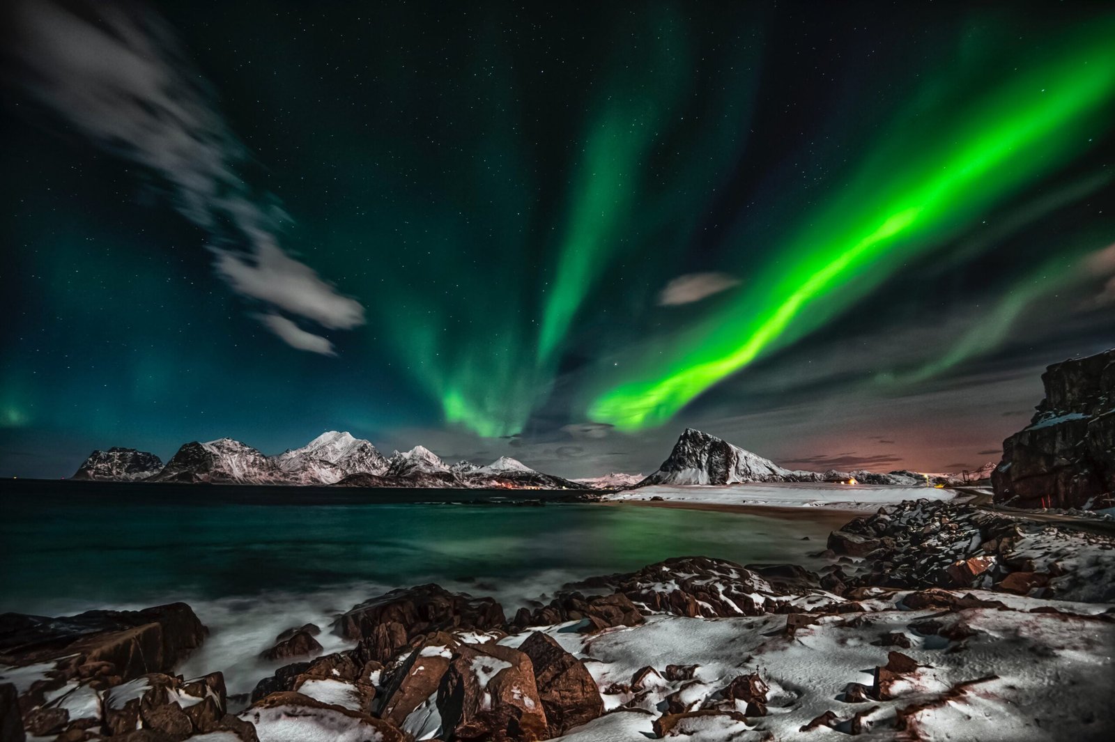 Vibrant green northern lights dancing over snowy mountain peaks, with rocky coastline and calm ocean waters in the foreground under a starry night sky.