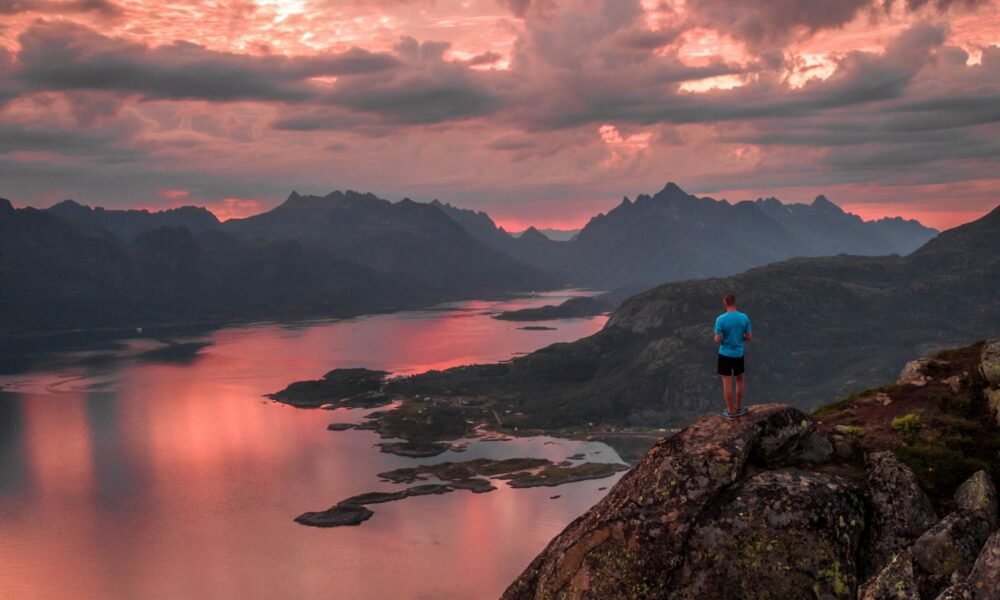 hiker overlooking a serene fjord in Norway at sunset with dramatic pink and orange clouds, rugged mountains in the distance, and shimmering reflections on calm waters.