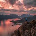 hiker overlooking a serene fjord in Norway at sunset with dramatic pink and orange clouds, rugged mountains in the distance, and shimmering reflections on calm waters.