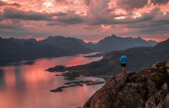 hiker overlooking a serene fjord in Norway at sunset with dramatic pink and orange clouds, rugged mountains in the distance, and shimmering reflections on calm waters.