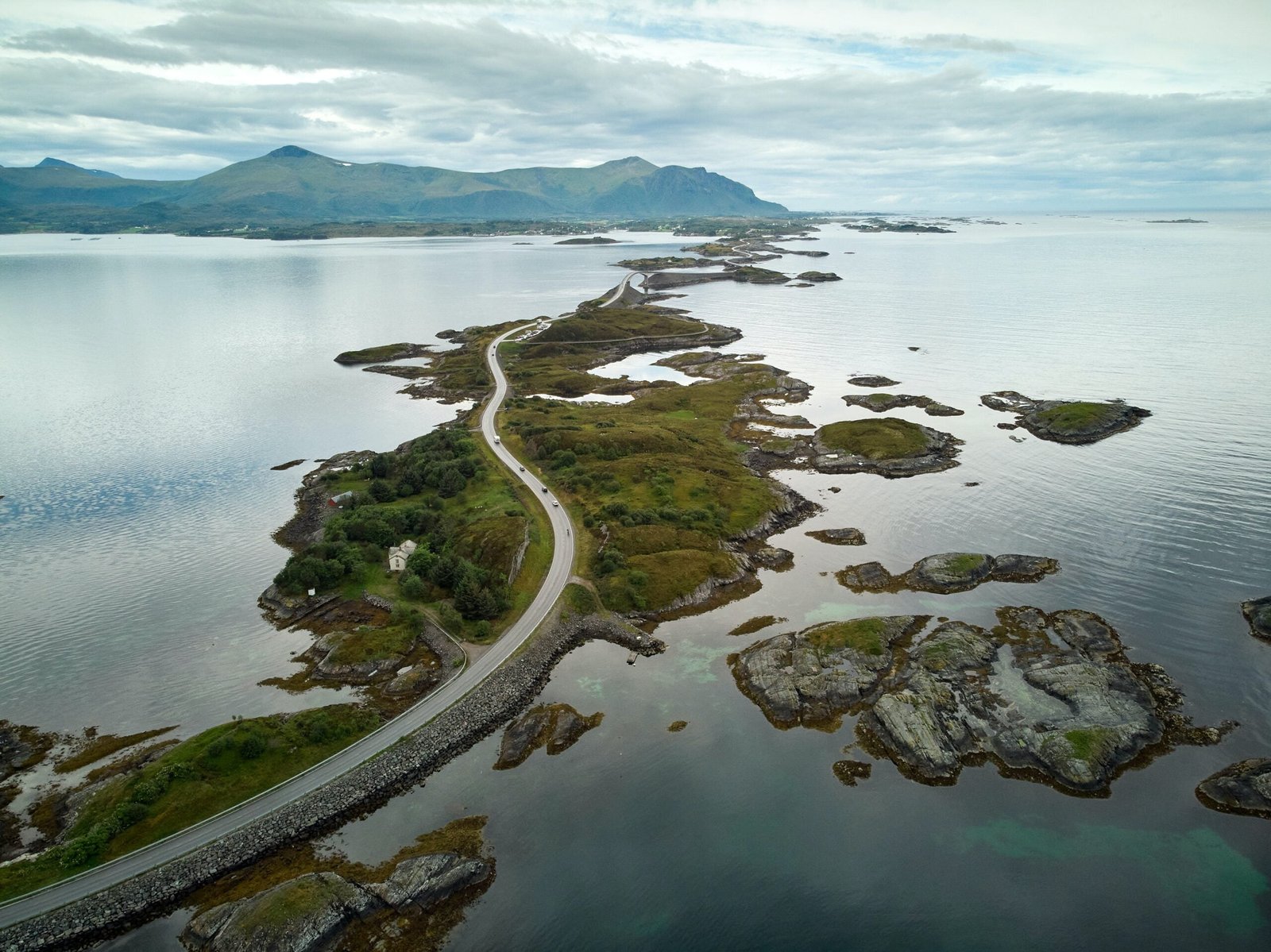 Aerial view of a winding coastal road amidst green Norwegian islets, with calm waters reflecting the overcast sky, and distant mountains on the horizon.