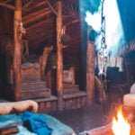 Rustic Viking-inspired interior with wooden carvings, fur-covered benches, a blue and white shield, and a central crackling fire emitting smoke under a timbered canopy.