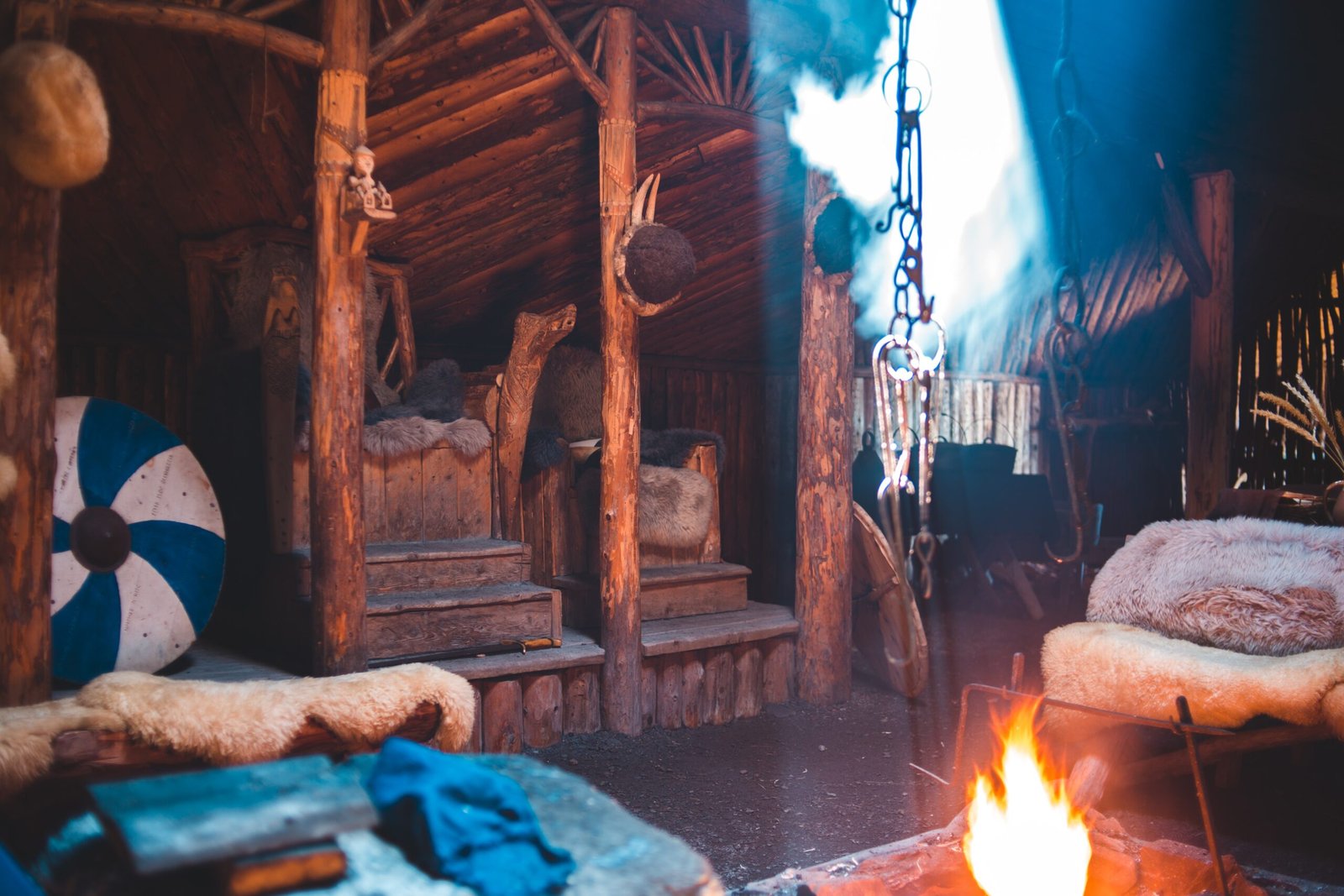 Rustic Viking-inspired interior with wooden carvings, fur-covered benches, a blue and white shield, and a central crackling fire emitting smoke under a timbered canopy.