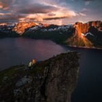 Person in yellow jacket sitting on a rugged cliff edge, overlooking a serene fjord with snow-capped mountains illuminated by sunset.