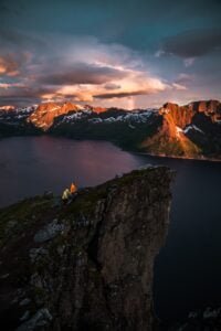 Person in yellow jacket sitting on a rugged cliff edge, overlooking a serene fjord with snow-capped mountains illuminated by sunset.