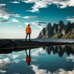 Man in orange jacket overlooking majestic Senja mountain range with reflection in water, serene Norwegian nature landscape for outdoor enthusiasts.