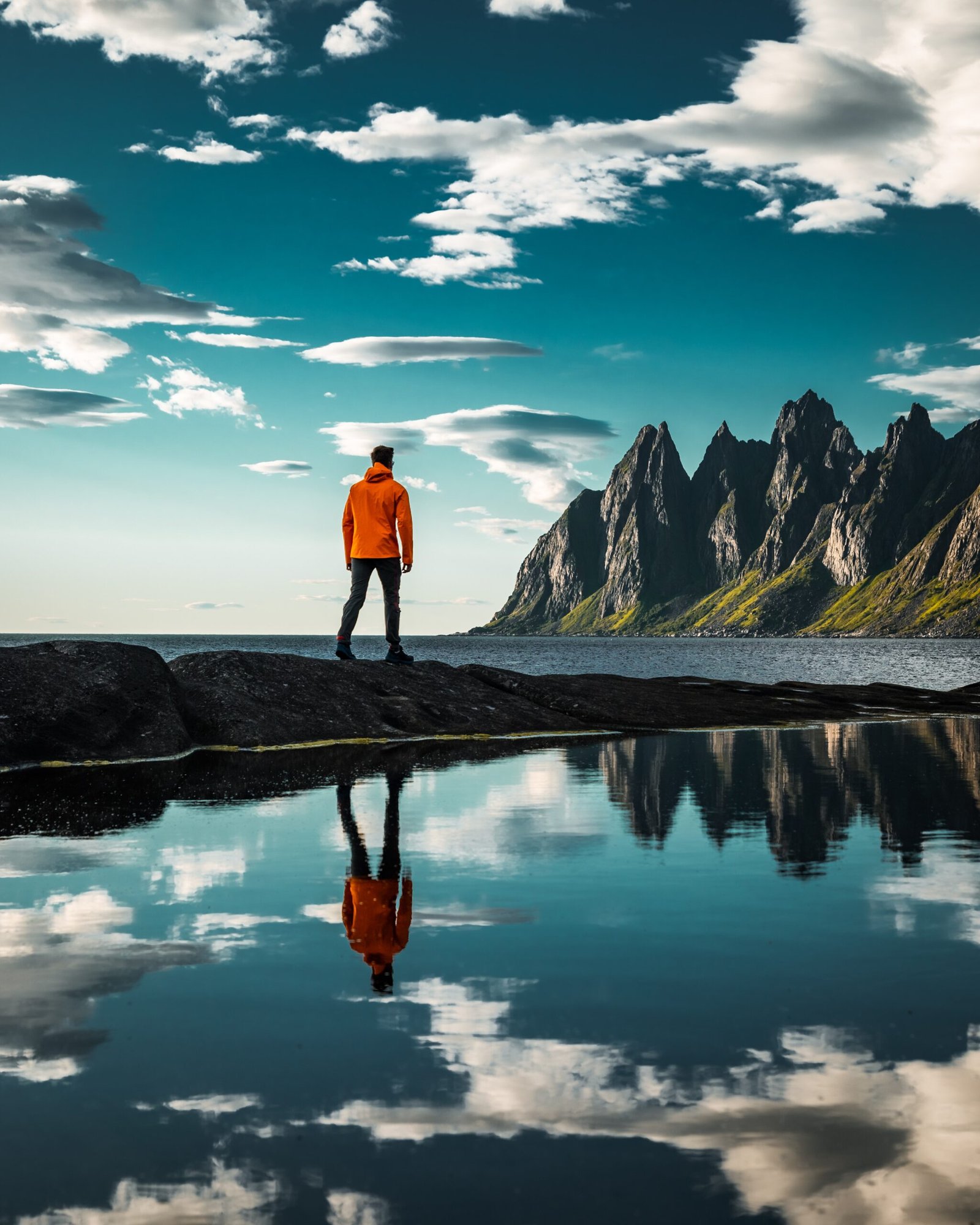 Man in orange jacket overlooking majestic Senja mountain range with reflection in water, serene Norwegian nature landscape for outdoor enthusiasts.